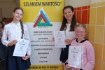 Daria Antolak, Alicja Franus i Bozhena Voloshyn na tle plakatu promocyjnego konkurs recytatorski „Wędrówki szlakiem wartości”.